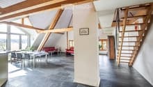 Open-plan living and dining area with exposed wooden beams, modern furniture including a red sofa, dining table with chairs, and a small stool.