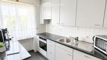 Modern kitchen with white cabinets, black countertops, stainless steel sink, oven, microwave, and a small table with a chair. A decorative rug with the word "KITCHEN" is on the floor.