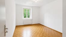 Empty room with wooden parquet flooring, white walls, a single window with white frame, radiator, and a door handle visible on the left side.