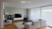 Modern living room with light-colored furniture, a large sectional sofa, ottoman, coffee table, TV, and plants.