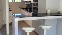 Modern kitchen with white cabinets, stainless steel appliances, black countertops, and two white bar stools at the island.