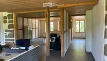 A modern, open-plan office space with wooden beams, a desk, chairs, and a cabinet. The room features a polished tile floor and large windows allowing natural light.