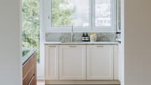 Modern kitchen with white cabinets, wooden flooring, and a large window overlooking greenery. Features a sink, faucet, and small decorative items on the counter.