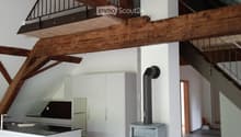 Modern kitchen area with white cabinets, a kitchen island, and a wood stove, featuring exposed wooden beams and a staircase leading to an upper level.
