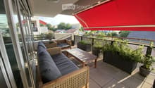 Balcony with a wooden sofa, a small table, a chair, and several potted plants. Features a red awning and a view of the surrounding area.