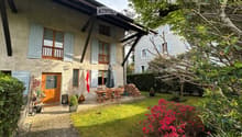 A well-maintained garden area with a wooden dining set, a closed umbrella, a stack of firewood, potted plants, and a flag. The garden is adjacent to a two-story house with light blue shutters and a wooden balcony.