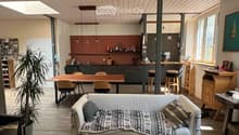 Open-plan living and dining area with a sofa, dining table, chairs, kitchen area, pendant lights, and a plant.