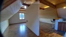 Bright, open attic space with wooden flooring, exposed wooden beams, and a small kitchen area featuring a counter and a stool.