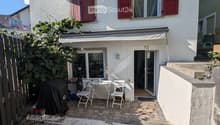 Small outdoor patio area with a table, chairs, and a covered grill, adjacent to a two-story house with red shutters and a balcony.