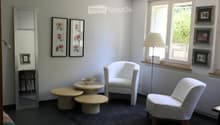 A modern, minimalist living space with white walls, dark flooring, and large windows. Features include a white armchair, small round tables, a floor lamp, and decorative picture frames.