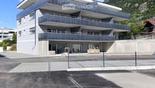 Modern three-story building with balconies, large windows, and a contemporary design, situated in a parking lot with a mountain backdrop.