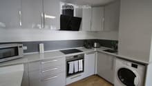 Modern kitchen with grey cabinets, white countertops, and stainless steel appliances including a microwave, coffee maker, toaster, oven, and washing machine. Features a range hood and a sink.