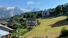 A picturesque village scene with a house featuring a balcony, surrounded by green lawns, trees, and snow-capped mountains in the background.