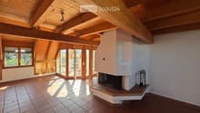 Bright, open room with exposed wooden beams, tiled flooring, large windows, and a fireplace. The space is empty, with no furniture or furnishings visible.