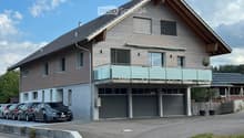 Two-story modern house with wooden siding, large windows, a balcony, and a garage with multiple parked cars.
