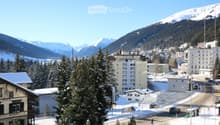 Panoramic view of a snowy town with mountains in the background, featuring buildings, trees, and a road.