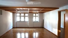 Empty room with wooden flooring, white walls, and large windows allowing natural light. The ceiling features exposed wooden beams, and there is a partially open wooden door leading to another space.