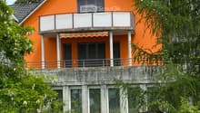 Two-story orange house with a raised ground floor, balcony, and surrounding greenery.