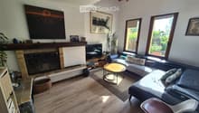Modern living room with wooden flooring, large windows, fireplace, sofa, coffee table, and decorative items.