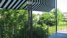 Balcony with metal railing, striped awning, and view of greenery and a distant church tower.
