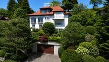 Two-story white house with red-tiled roof, multiple windows, balconies, and a garage surrounded by lush greenery and trees.