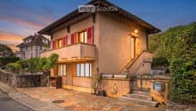 Two-story house with beige facade, red shutters, and a raised ground floor accessed by stairs. Features a stone-paved entrance area, small garden, and a balcony on the upper level.