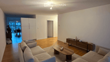 Modern living room with light-colored wooden flooring, a large sectional sofa, a coffee table, and a side table with decorative items. The room features white walls and a ceiling light fixture.