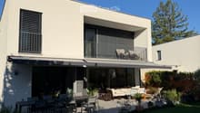 Modern house exterior with a spacious patio area featuring a dining table, chairs, a sofa, and potted plants. The house has large windows and a balcony with additional seating.