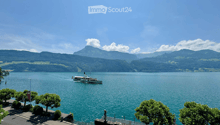 A scenic view of a lake with a boat sailing on it, surrounded by lush green trees and distant mountains under a clear blue sky.