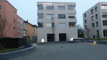 Modern multi-story building with large windows and balconies, surrounded by paved area and low lighting.