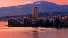 A scenic view of a cityscape at sunset, featuring a body of water in the foreground, with buildings and trees lining the shore, and mountains in the background.