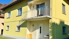 Two-story yellow house with white trim, multiple windows, a balcony on the upper floor, and a small patio area with a table and chairs.