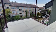 Balcony with metal railing, concrete floor, overlooking a residential area with buildings and trees.