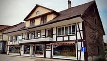 Two-story building with a tiled roof, multiple windows, and a storefront with awnings and signage. The ground floor has glass windows displaying various items, and the upper floor features balconies and decorative railings.