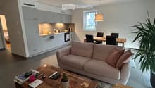 A modern, open-concept living and dining area with a light pink sofa, wooden coffee table, dining table with chairs, and a small kitchen area with white cabinets and stainless steel appliances.