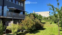 Lush green garden with well-maintained lawn, various shrubs, and trees, featuring a modern multi-story building with balconies and large windows in the background.