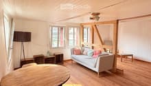Bright living room with wooden floors, white paneled walls, and large windows. Features a light grey sofa with colorful cushions, a round wooden table, and a tall floor lamp.