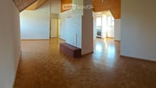 Empty room with wooden flooring, white walls, and a slanted wooden ceiling. A small bench is placed against one wall, and a doorway leads to another room with a visible kitchen area.