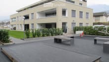 Modern two-story building with beige facade, multiple windows, balconies, and a landscaped courtyard with benches and greenery.