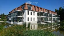 Modern two-story building with white walls, red-tiled roof, and large glass balconies overlooking a serene water body.