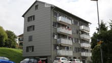Four-story apartment building with grey facade, multiple balconies, and flower boxes. Several cars and a motorcycle are parked in front of the building.