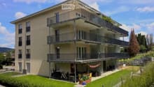 Modern three-story apartment building with multiple balconies, surrounded by greenery and a small garden area with outdoor furniture.