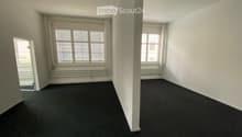 Empty room with white walls, dark carpet, large windows with blinds, and a radiator under one window.