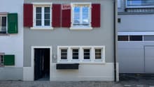 Two-story house with light gray walls, red shutters, and a red balcony railing. The ground floor has a black door and several windows, while the upper floor features additional windows and a small balcony. The roof is tiled with brown tiles and has dormer