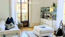 Elegant living room with a white sofa, ottoman, glass coffee table, bookshelf, and a chandelier hanging from the ceiling. The room features wooden flooring and large windows with sheer curtains, leading to a balcony.