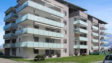 Modern multi-story apartment building with glass balconies, light-colored facade, and surrounding green lawn.