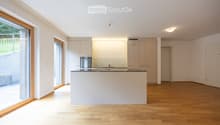 Modern, minimalist kitchen with white cabinetry, stainless steel appliances, and a central island. Large windows provide natural light, and the floor is made of light wood.