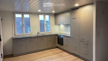 Modern kitchen with light grey cabinets, wooden flooring, and large windows offering a view of the roof.