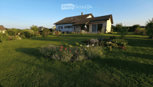 A well-maintained garden with a lawn, flower beds, and a house in the background. The house has a tiled roof, large windows, and a covered patio area with outdoor furniture.