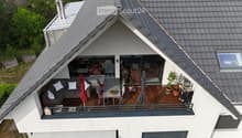 Wooden balcony with grey sofa, dining table, chairs, umbrella, and potted plants.
